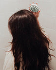 A woman with long wavy brown hair using a sage green scalp massager to massage the back of her head.