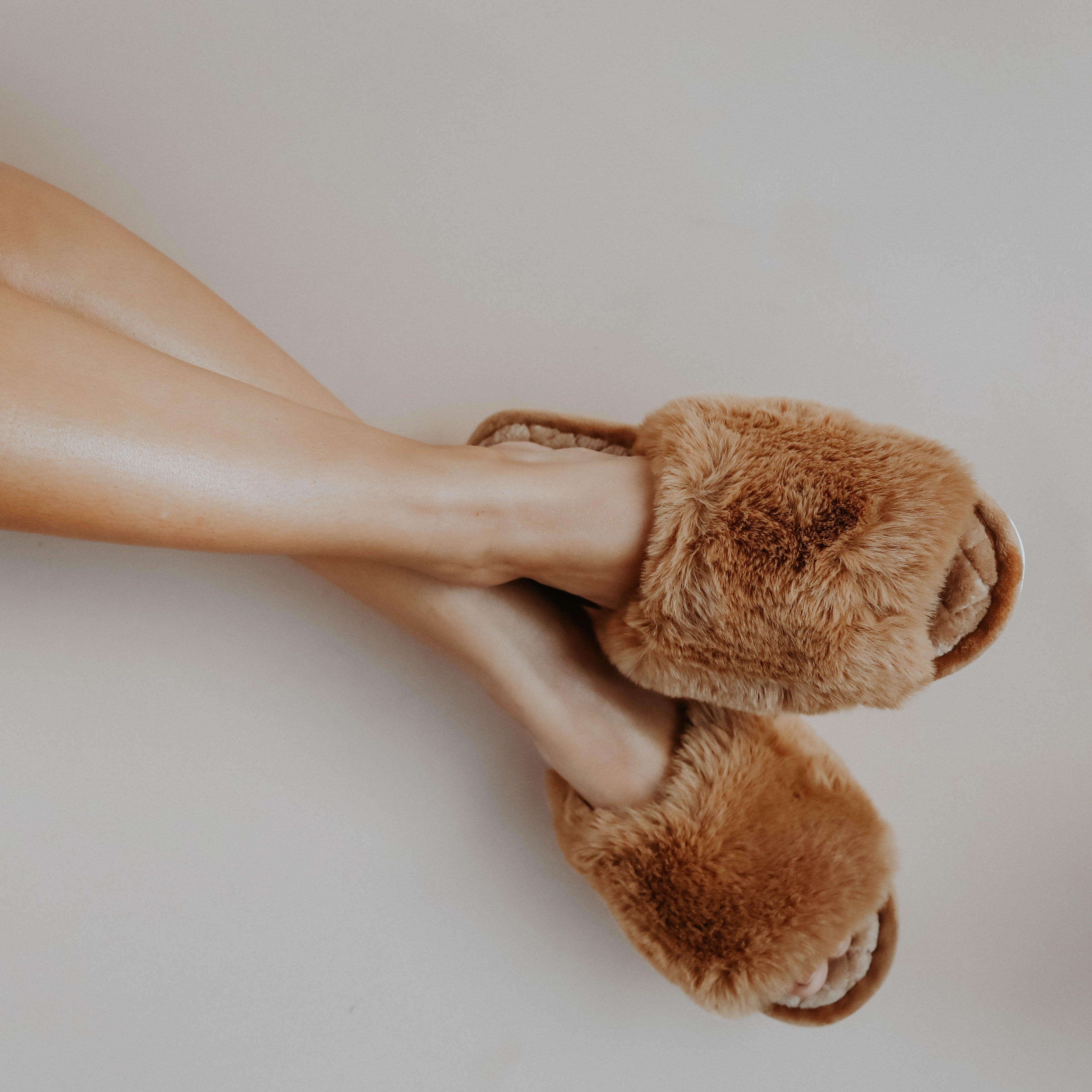 A girl crossing her legs wearing brown fuzzy slippers against a white background