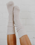 A girl with tan legs wearing mid-calf stone grey fuzzy socks with her legs up in the air criss-crossing against a white background