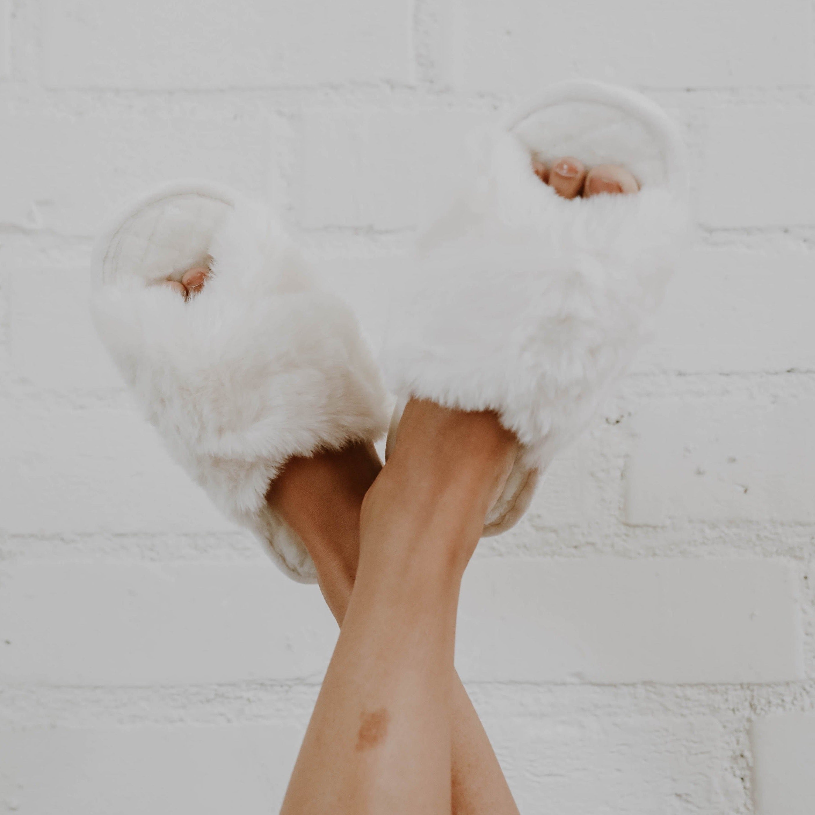A girl crossing her legs in the air wearing white fuzzy slippers against a white background