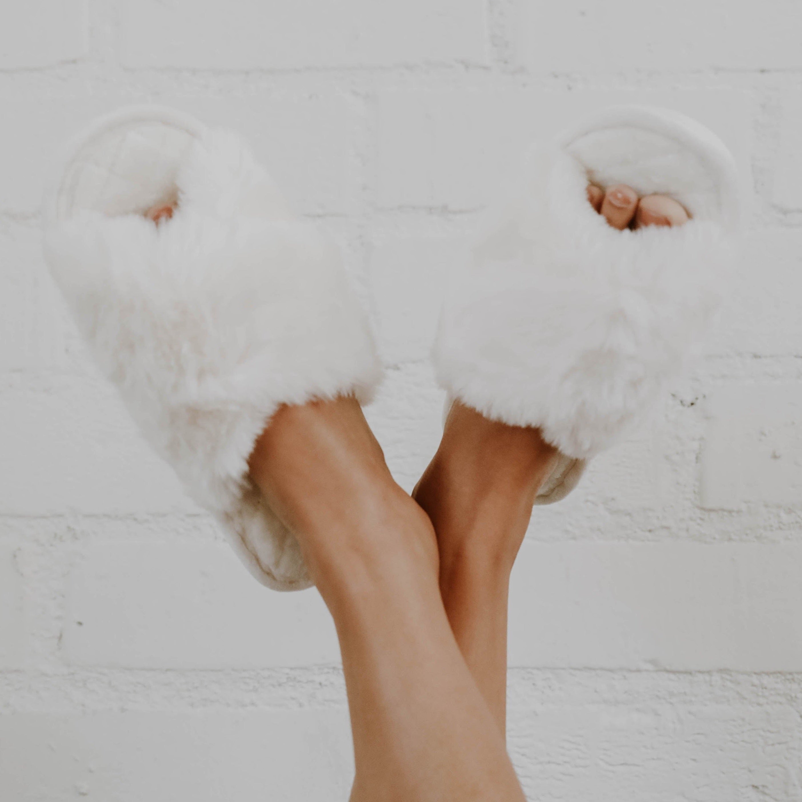 A girl crossing her legs in the air wearing white fuzzy slippers against a white background