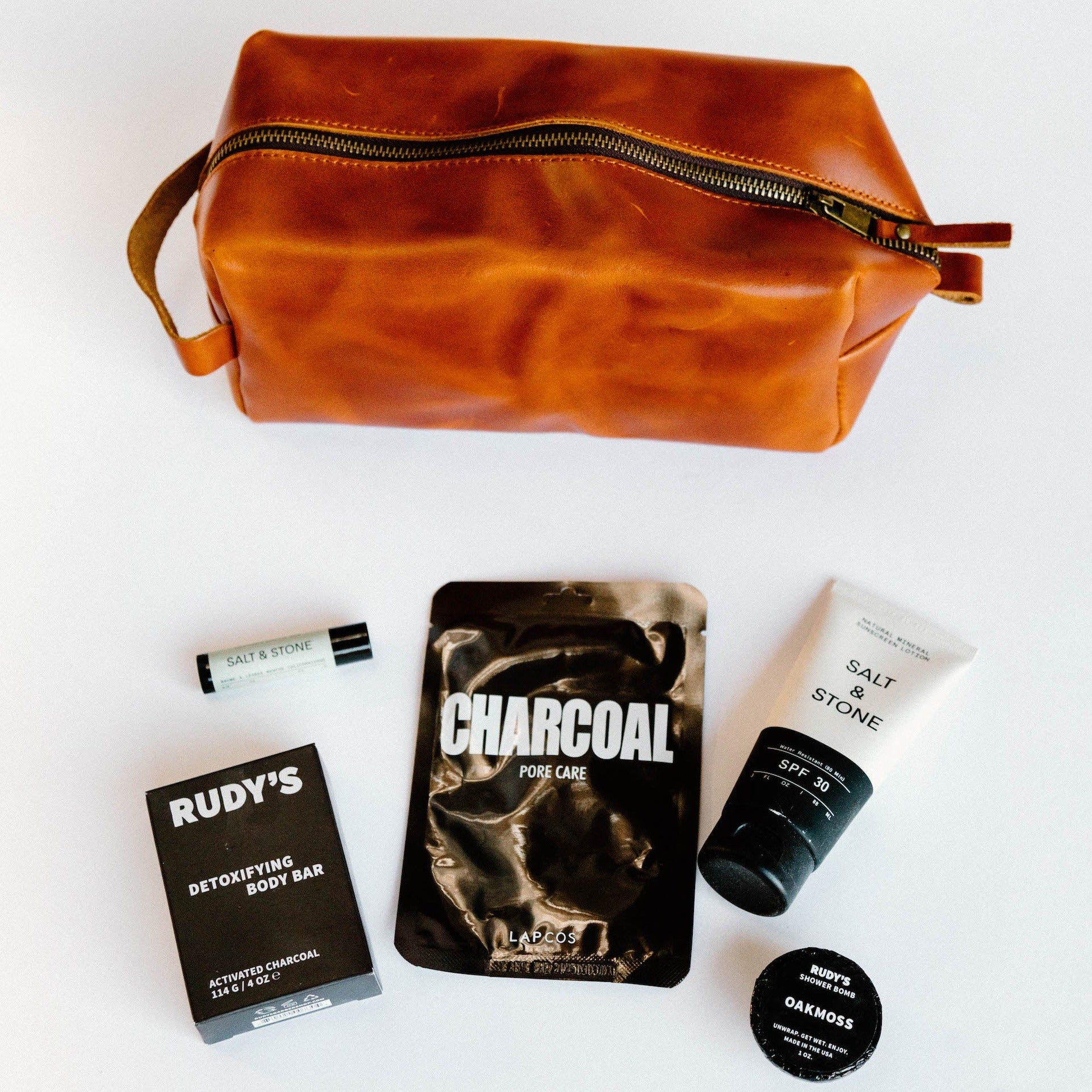 A brown leather dopp kit with various black toiletries laying against a white background.
