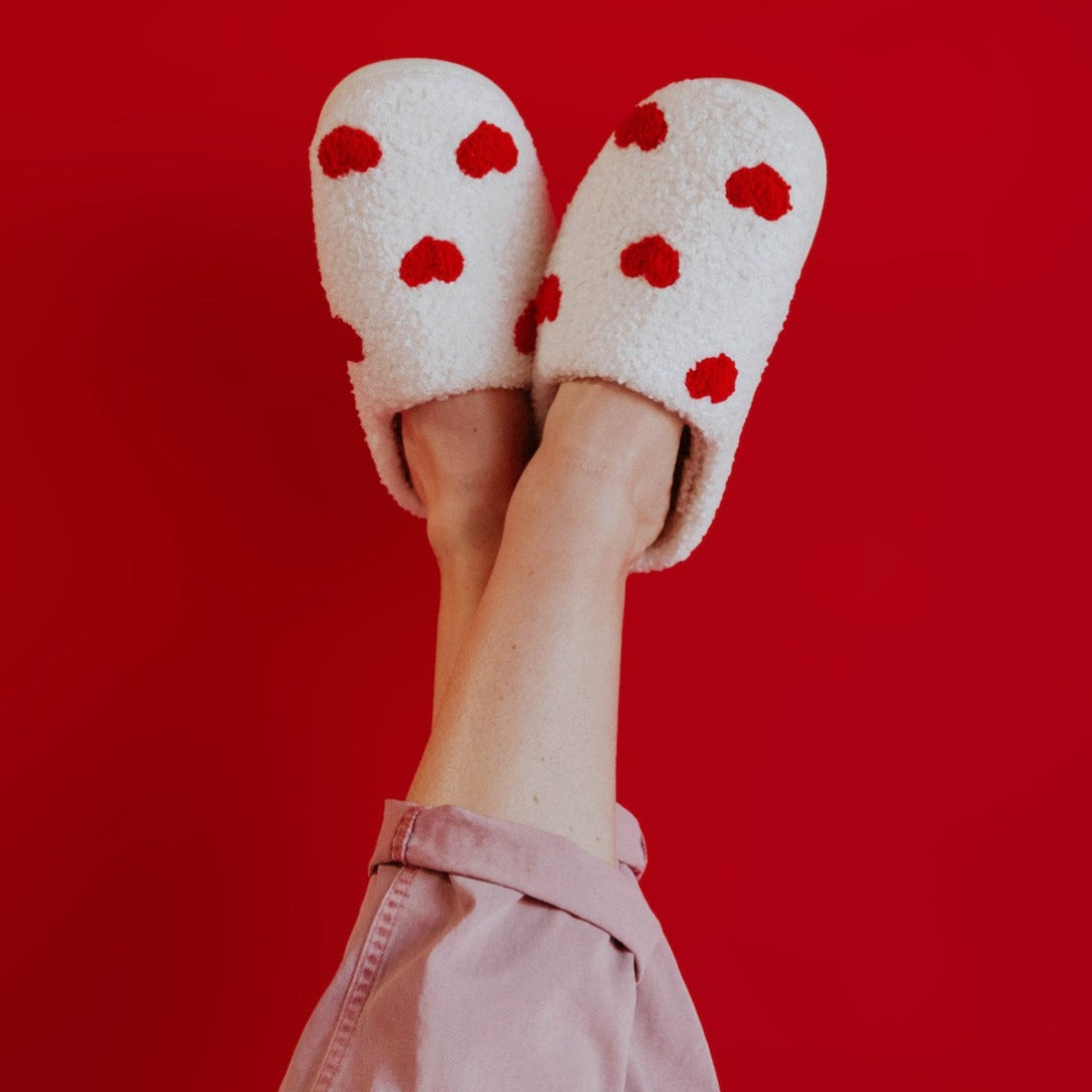 a woman's legs in the air wearing a pair of white slippers that have red hearts on them. her legs are against a red background.