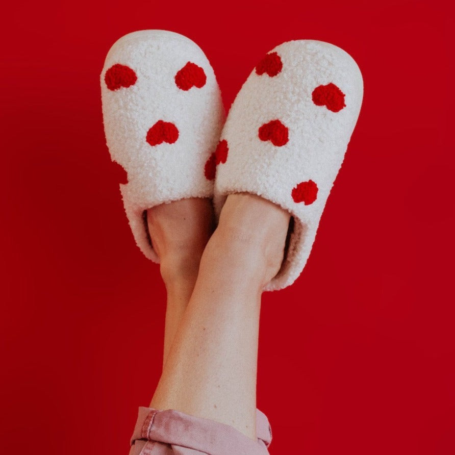 a woman&#39;s legs in the air wearing a pair of white slippers that have red hearts on them. her legs are against a red background.
