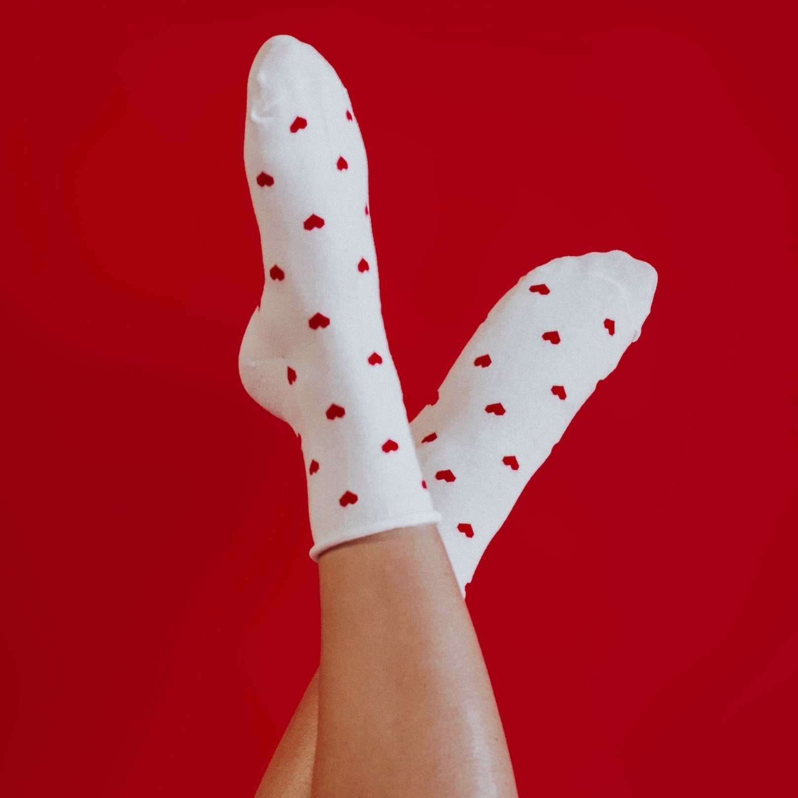 a woman's legs in the air wearing a pair of white crew socks that have tiny red hearts on them. her legs are against a red background.