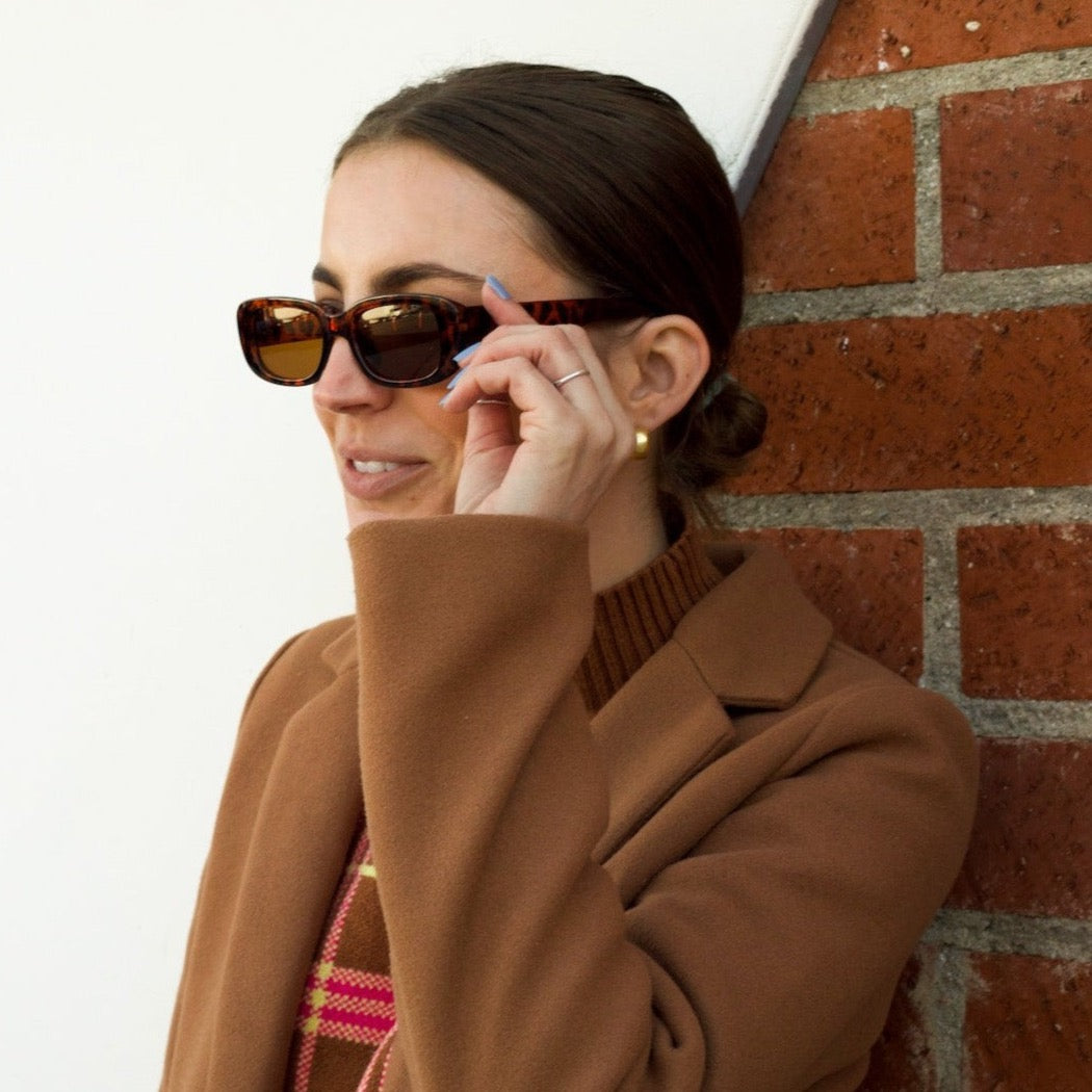 A girl with brown hair in a low bun wearing a brown sweater and brown long wool coat standing in front of a white and brick background wearing brown tortoise rectangle sunglasses.
