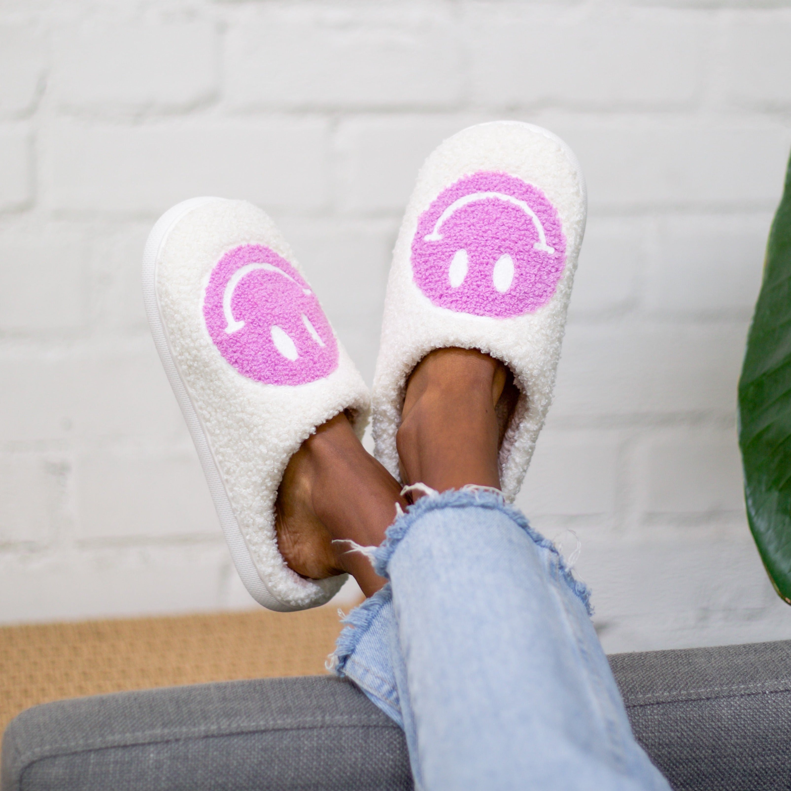 A girl wearing white slippers with orchid smiley faces on them. Her legs are up against a white brick background.