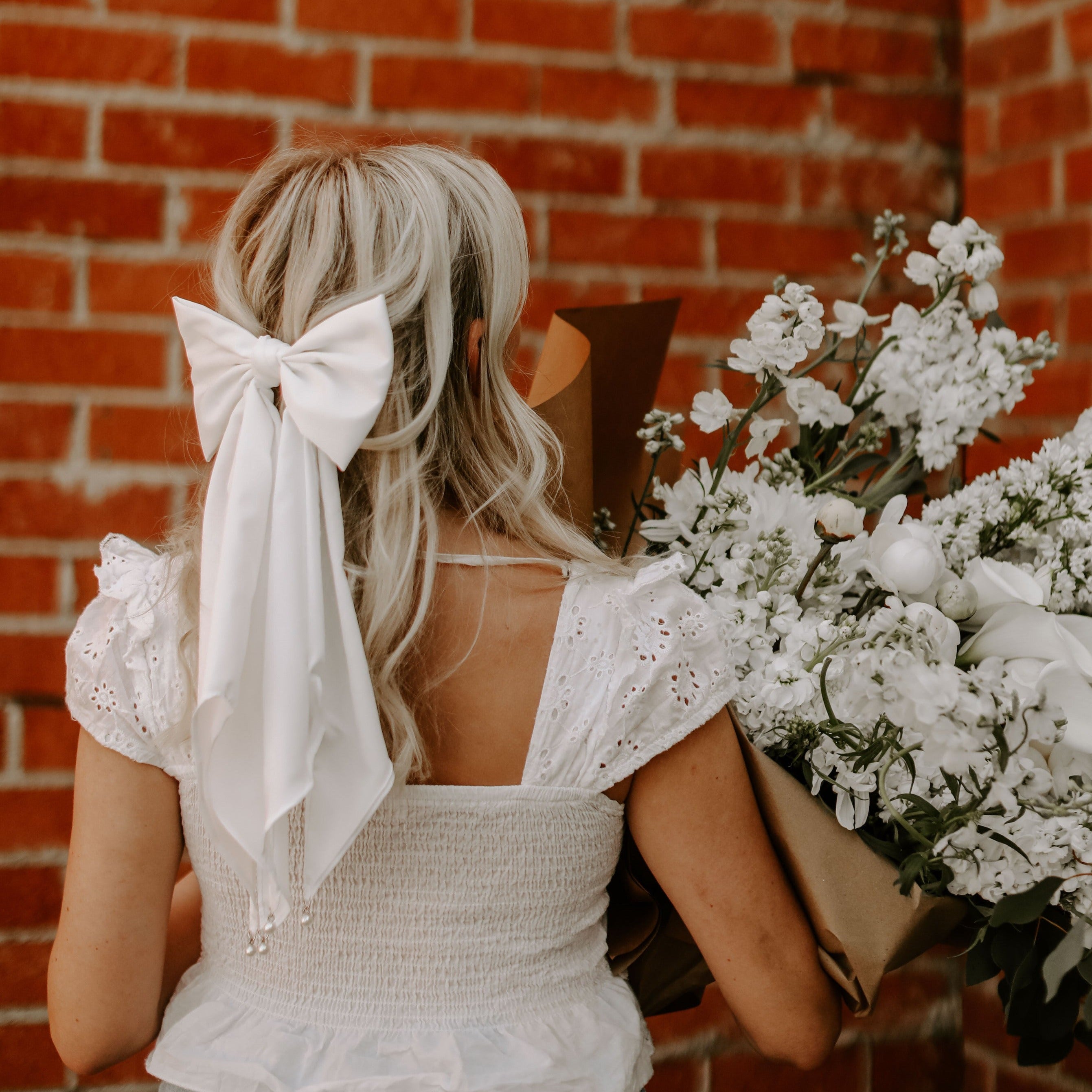 Blonde female facing brick wall with long bridal bow tying hair back half up , half down wearing white shirt and holding bundle of white flowers.