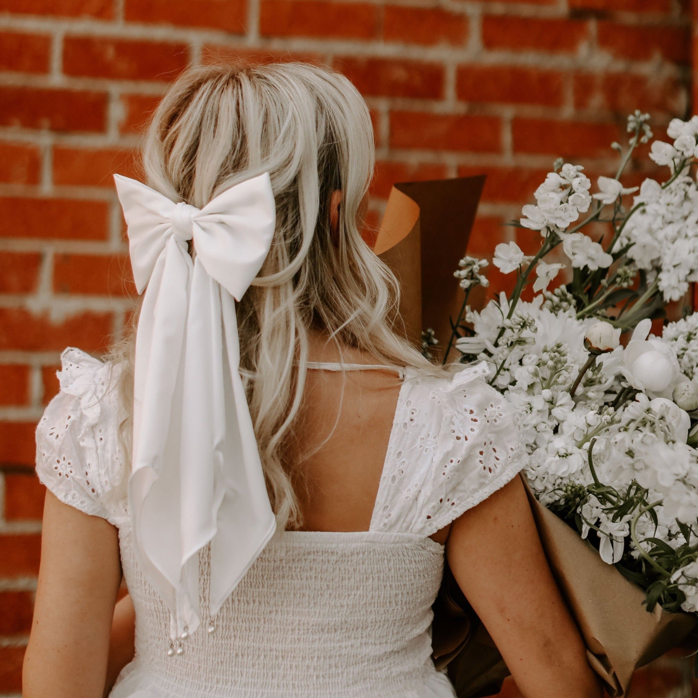 Blonde female facing brick wall with long bridal bow tying hair back half up , half down wearing white shirt and holding bundle of white flowers.