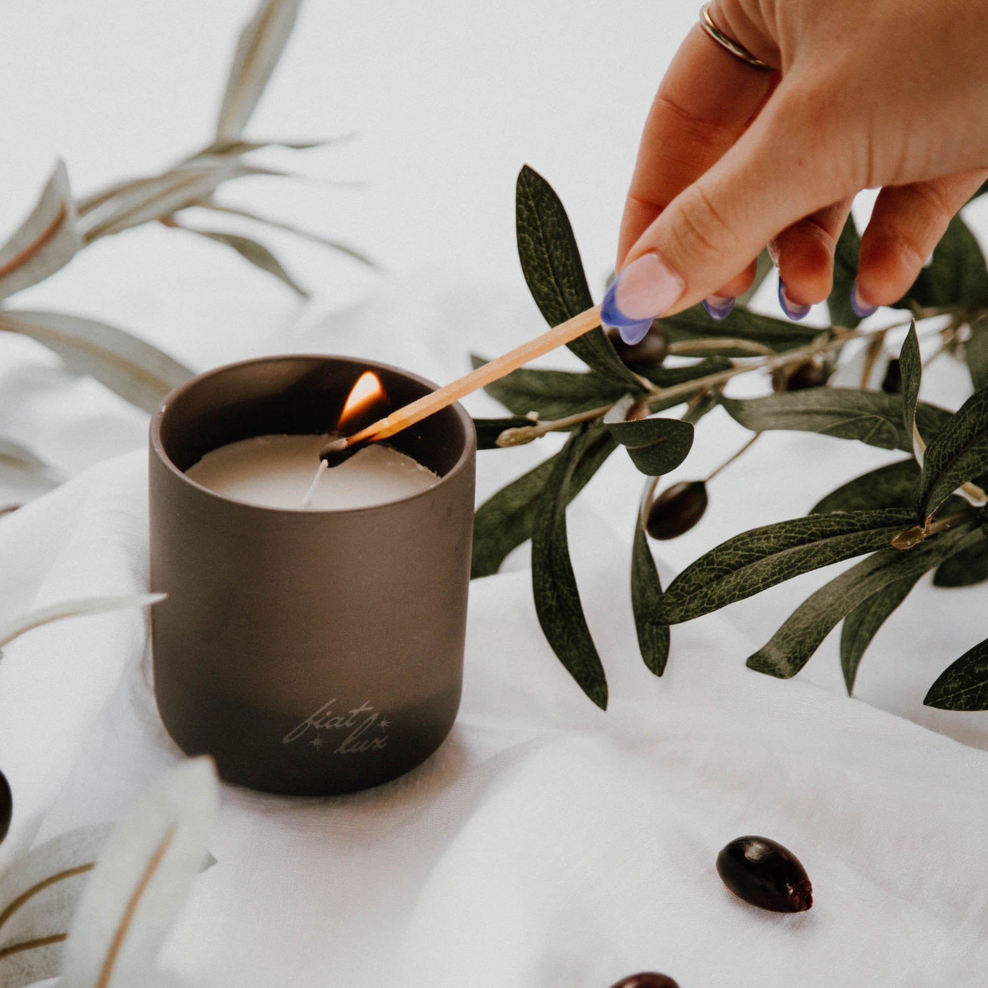 A black ceramic candle jar filled with white wax and a lit wick (Fiat Lux Valldemossa) surrounded by olive branches and Kalamata olives.