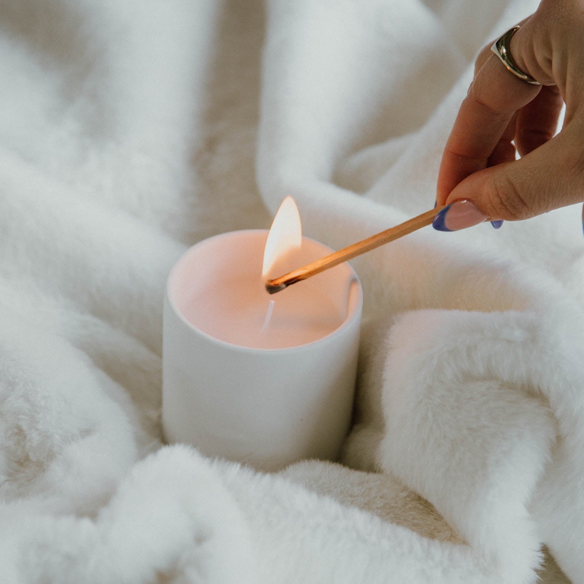 A white ceramic candle jar filled with white wax and a lit wick (Fiat Lux Copenhagen) surrounded by a fluffy white blanket.
