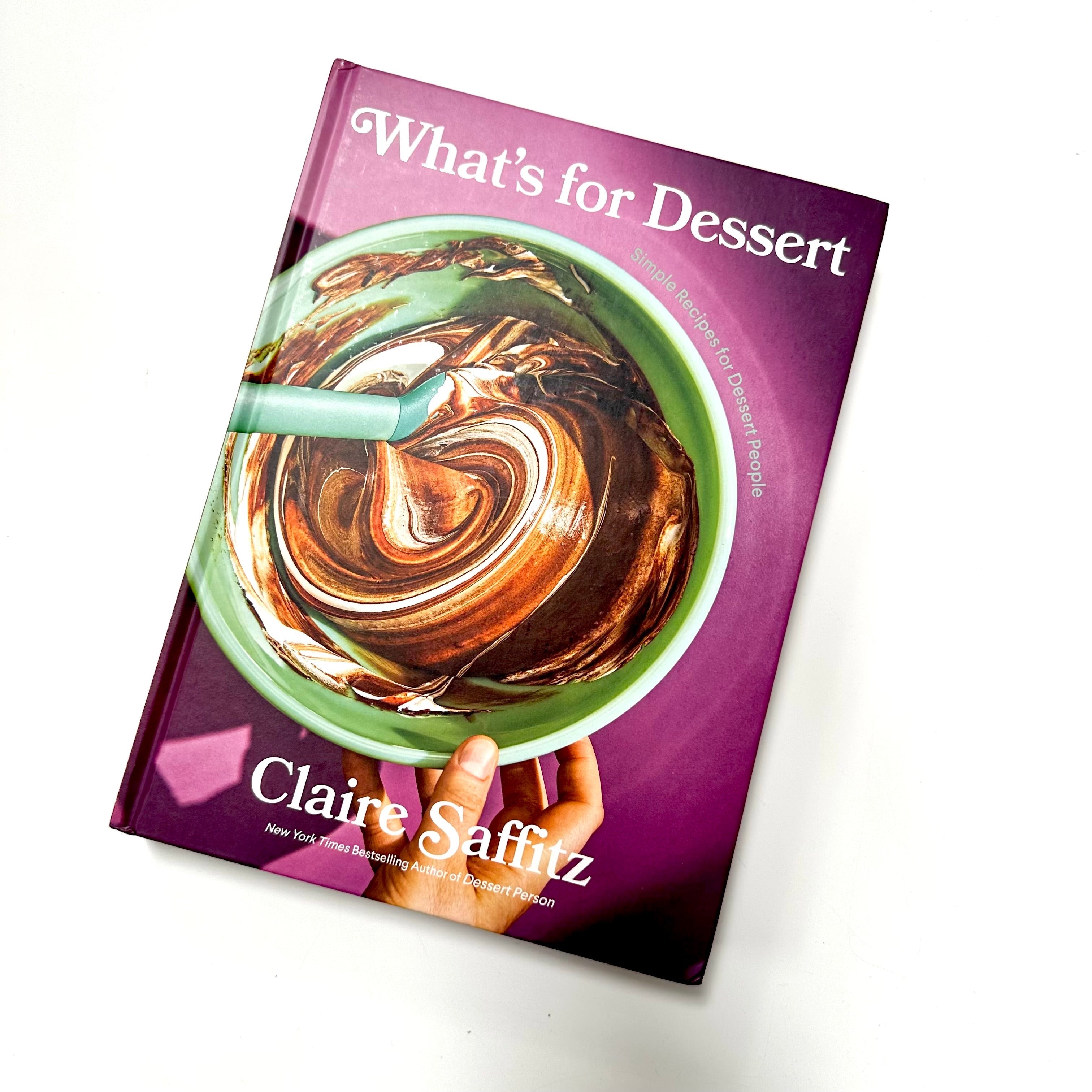 A purple hardcover book with a mixing bowl of brown mixture. White text on book reads, &quot;What&#39;s for Dessert Simple Recipes for Dessert People Claire Saffitz&quot;. Photographed on white background.