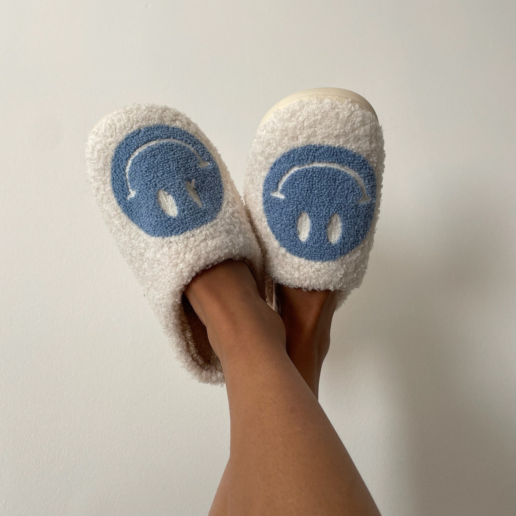A girl wearing white slippers with blue smiley faces on them. Herr legs are up against a white background