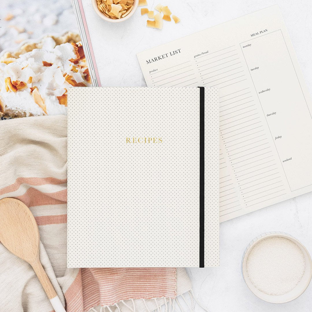 White recipe book with black polka dots and gold foil writing sits on a tea towel, market list, cook book wooden spoon and ramekin. 