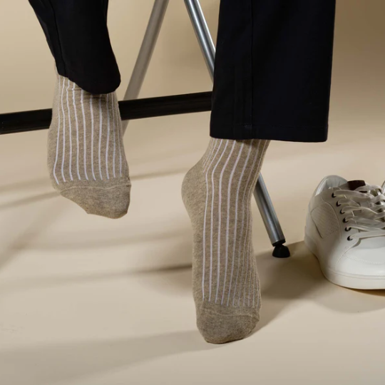 Men's feet wearing beige striped socks and black pants photographed in front of a tan backdrop with metal chair legs and white shoe in background