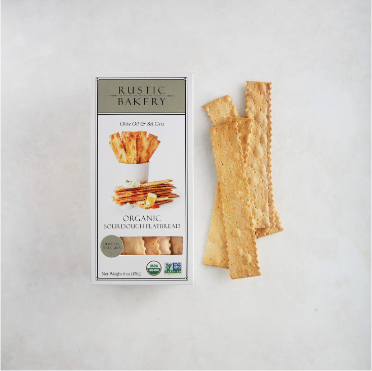 A box of the Rustic Bakery Olive Oil &amp; Sel Gris Organic Sourdough Flatbreads laying to the right of 4 flatbread crackers. Photographed on light grey background.