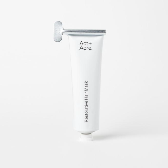 A white squeeze tube with silver rolling dispenser at tup. Black text on tube reads, &quot;Act+Acre Restorative Hair Mask&quot;. Photographed on white background. 