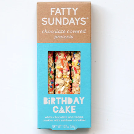 A rectangular paper box containing white chocolate and sprinkle covered pretzel sticks. Packaging is light brown and blue with white text that reads, "FAtTY SUNDAYS chocolate covered pretzels Birthday Cake white chocolate and vanilla cookies with rainbow sprinkles". Photographed on white background.