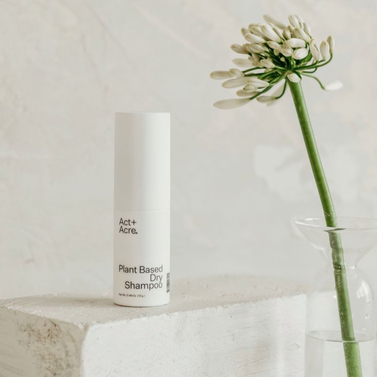 A cylindrical, white plastic bottle that reads, &quot; Act + Acre Plant Based Dry Shampoo&quot; in black text sitting on top of white stone block with white flower in clear glass vase on the right. Photographed on white background.