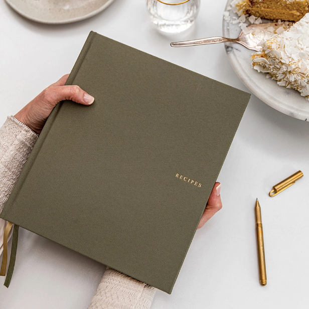 A Caucasian woman's hands hold olive green linen journal with "RECIPES" embossed in gold on center right over white table that also holds uncapped gold pen, marble platter with cake and silver serving knife and glass cup. 