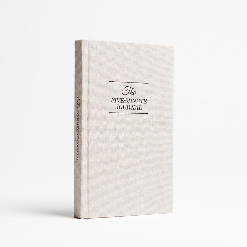 A cream, linen covered, hard cover journal with black embossed writing that reads, &quot;The Five Minute Journal&quot; on front and along spine photographed on white background.