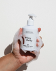 A caucasian hand covered in soap suds holds up a white plastic bottle with pump nozzle in front of white background.