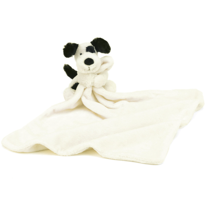 A black and cream stuffed animal dog with arms wrapped around one corner of cream soother blanked photographed on white background.