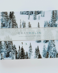 A rectangular puzzle box with photograph of snow covered ski slope and pink trees all over. Rectangular beige ribbon across middle of box reads, "Gray Malin Double-Sided Puzzle | 500 Pieces". Photographed on white background.