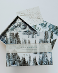 Gray Malin Winter Holiday Puzzle Box open showing loose pieces in bottom of box and images of completed puzzle (skiers on white, snow covered landscape and snow covered ski slope with pine trees). Photographed on white background.