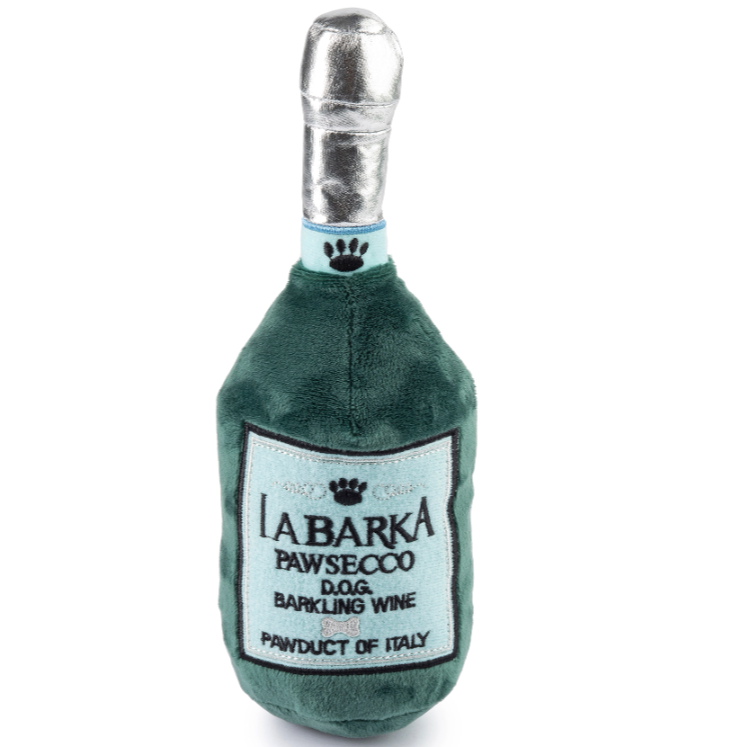 A plush dog toy shaped like a dark blue bottle of prosecco with metallic fabric top. Team label on front has black embroidered writing reading, "LaBarka Pawsecco D.O.G. barkling Wine Pawduct of Italy". Photographed on white background.