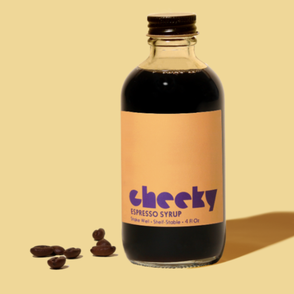 A glass bottle with black metal screw cap and orange label filled with dark brown espresso syrup. Purple text on bottle reads, "Cheeky Espresso Syrup Shake Well".  Photographed on light yellow background with espresso beans in foreground.