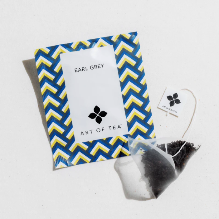 a clear tea sachet with white Art of Tea Label next to tea bag packaging with blue and yellow geometric design