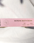 A long, pink, rectangular box with gold writing that reads, "Ollie Blossom Serene Bath Soak Bath Notes Peaceful, Floral, Tranquil" photographed on white linen