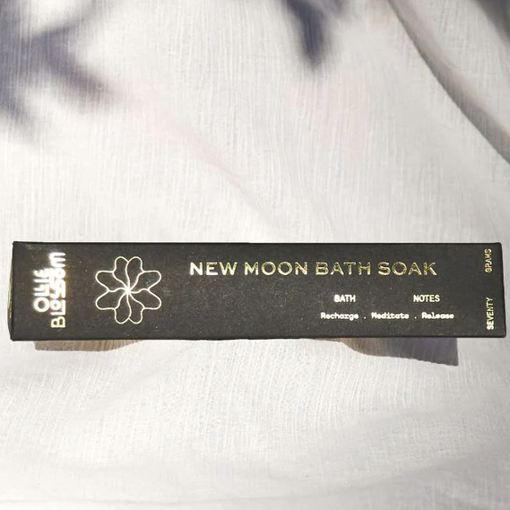 A black rectangular box with gold writing that reads, "Ollie Blossom New Moon Bath Soak Bath Notes Recharge, Meditate, Release" photographed on white linen.