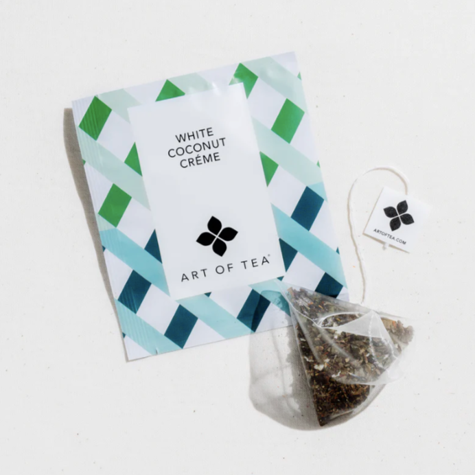 A white mesh tea sachet filled with loose, ground tea next to white paper packaging with green and blue lattice design and black text that reads, "White Coconut Crème Art of Tea" photographed on white background