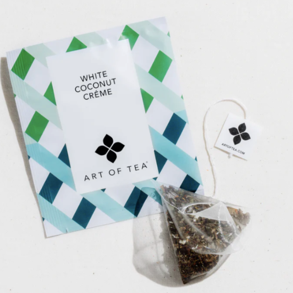 A white mesh tea sachet filled with loose, ground tea next to white paper packaging with green and blue lattice design and black text that reads, "White Coconut Crème Art of Tea" photographed on white background