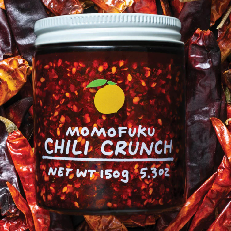 Image shows Momofuku's Chili Crunch in a clear jar with white lid, lying on a bed of chiles. 