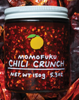 Image shows Momofuku's Chili Crunch in a clear jar with white lid, lying on a bed of chiles. 