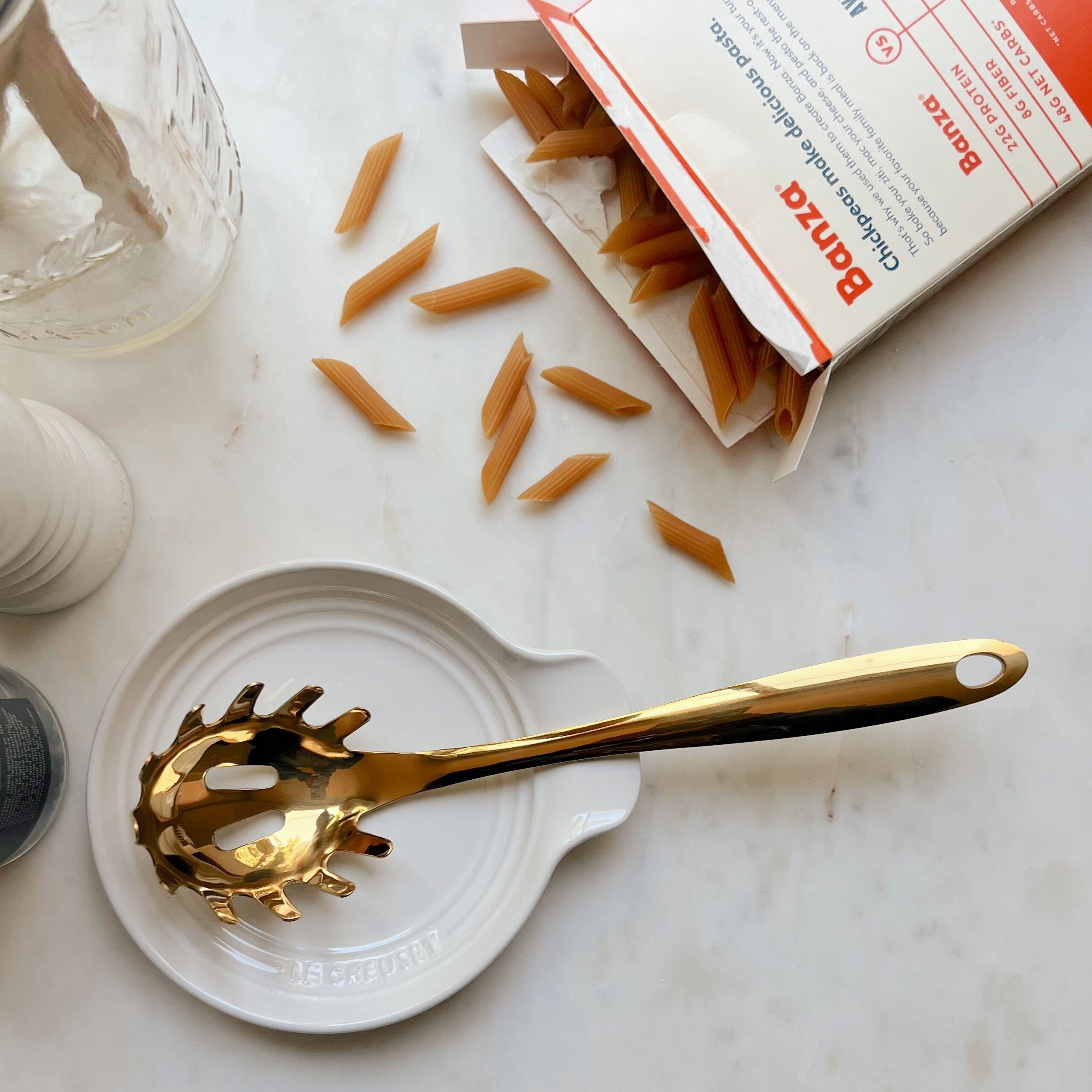 A gold pasta spoon laying on a white ceramic Le Creuset spoon rest next to Banza pasta noodles spilling out of their package on a white marble counter.