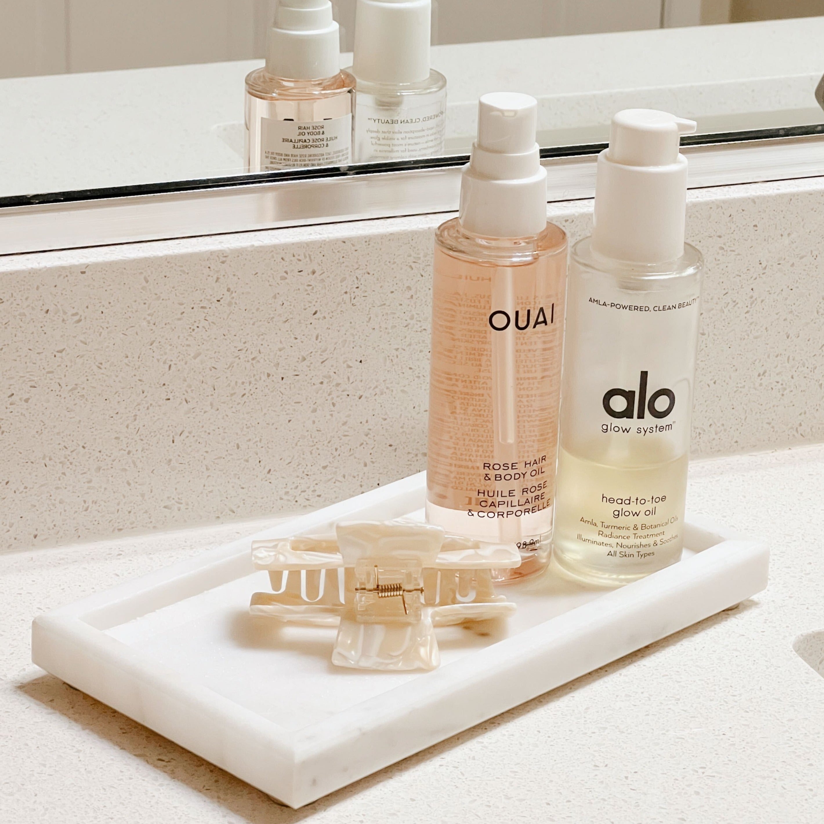 A white marble tray sitting on a white bathroom counter holding a white marble hair clip, Ouai rose hair & body oil, and alo head-to-toe glow oil.