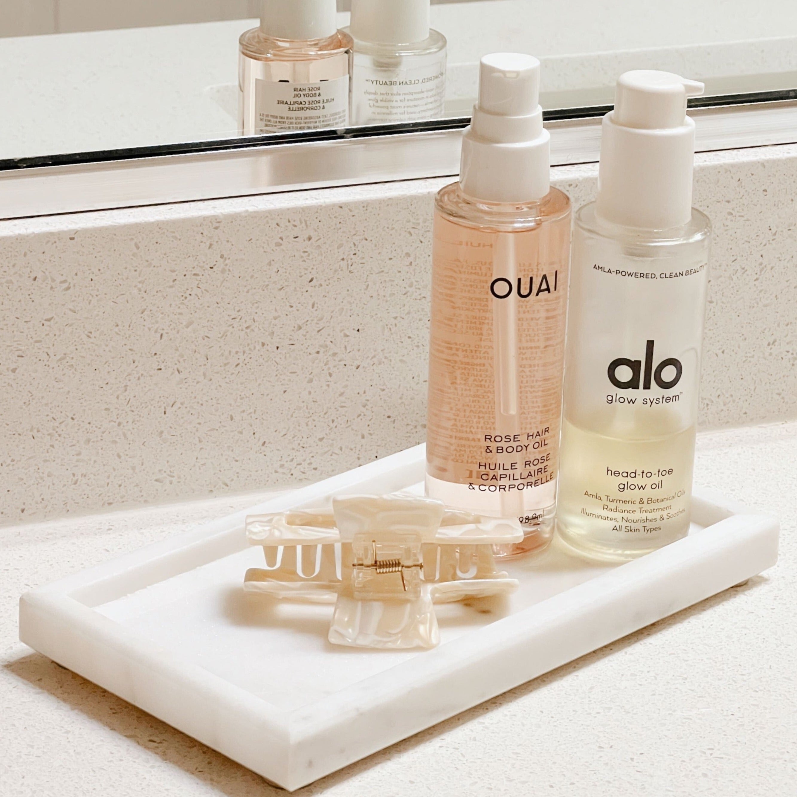 A white marble tray sitting on a white bathroom counter holding a white marble hair clip, Ouai rose hair & body oil, and alo head-to-toe glow oil.