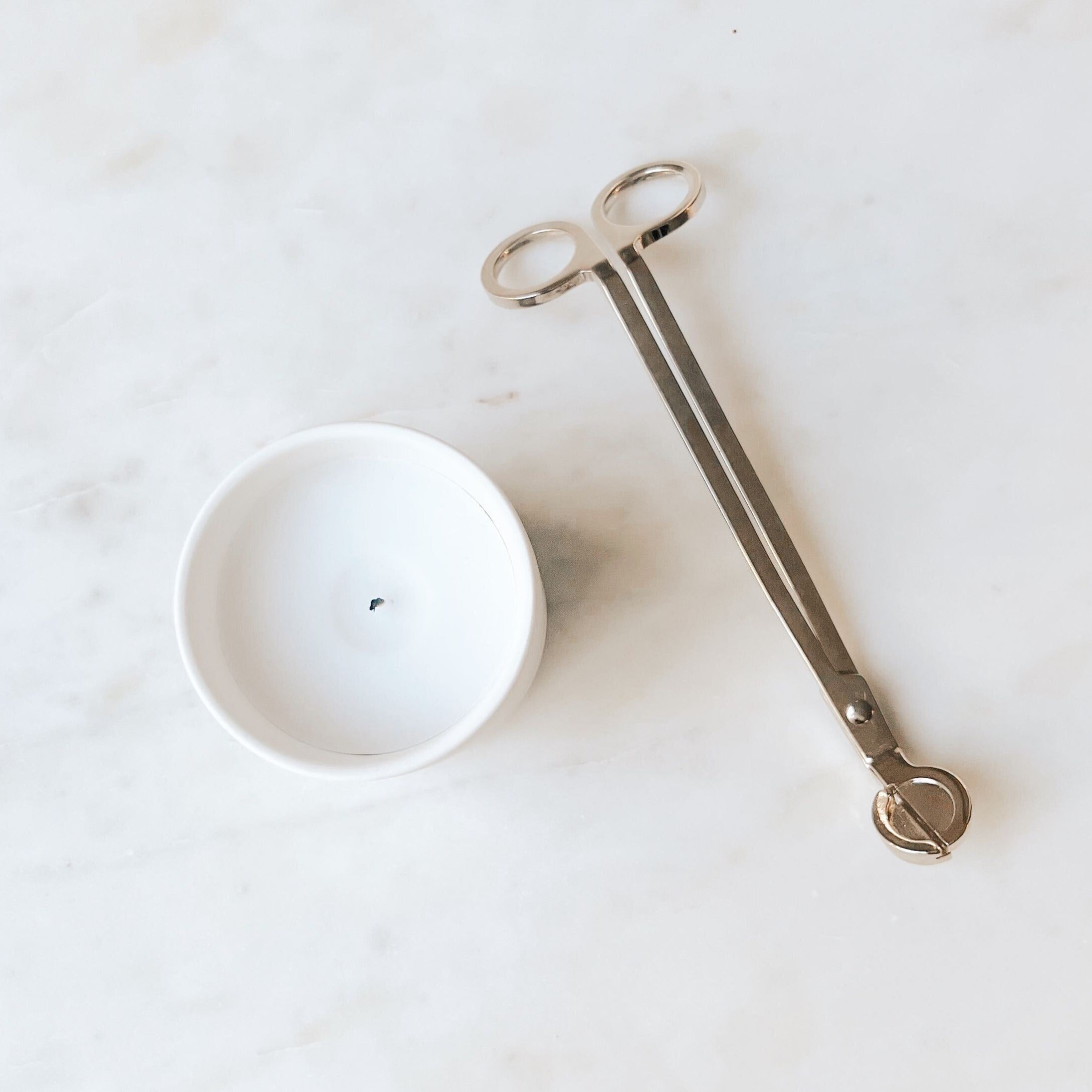 A gold wick trimmer laying next to a white Fiat Lux Copenhagen candle on a white marble table.