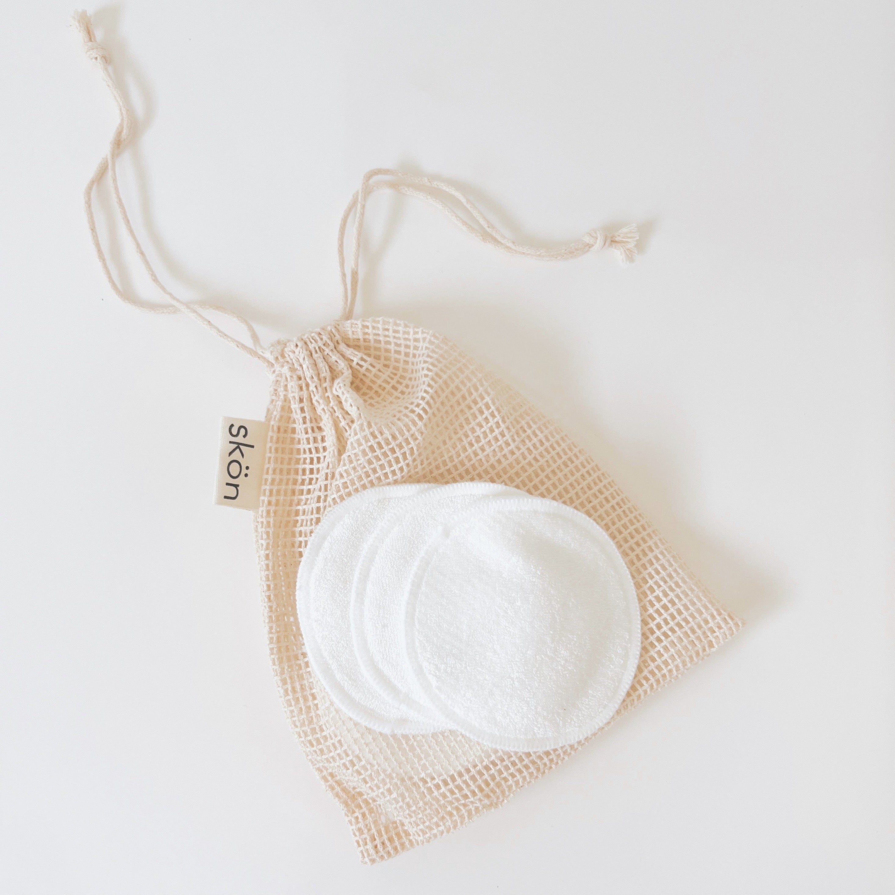 8 reusable bamboo cotton makeup rounds laying on a tan colored mesh bag against a white marble background
