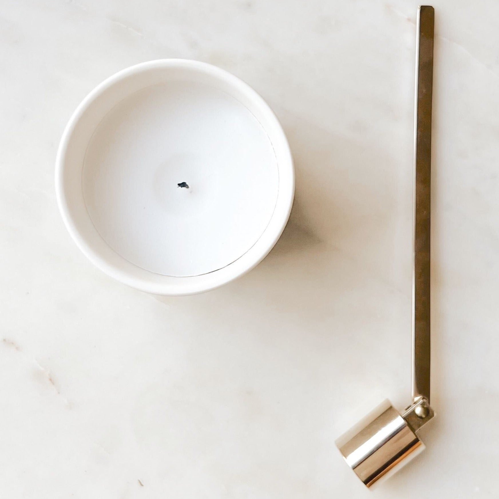 A gold candle snuffer laying next to a white Fiat Lux Copenhagen candle on a white marble table.