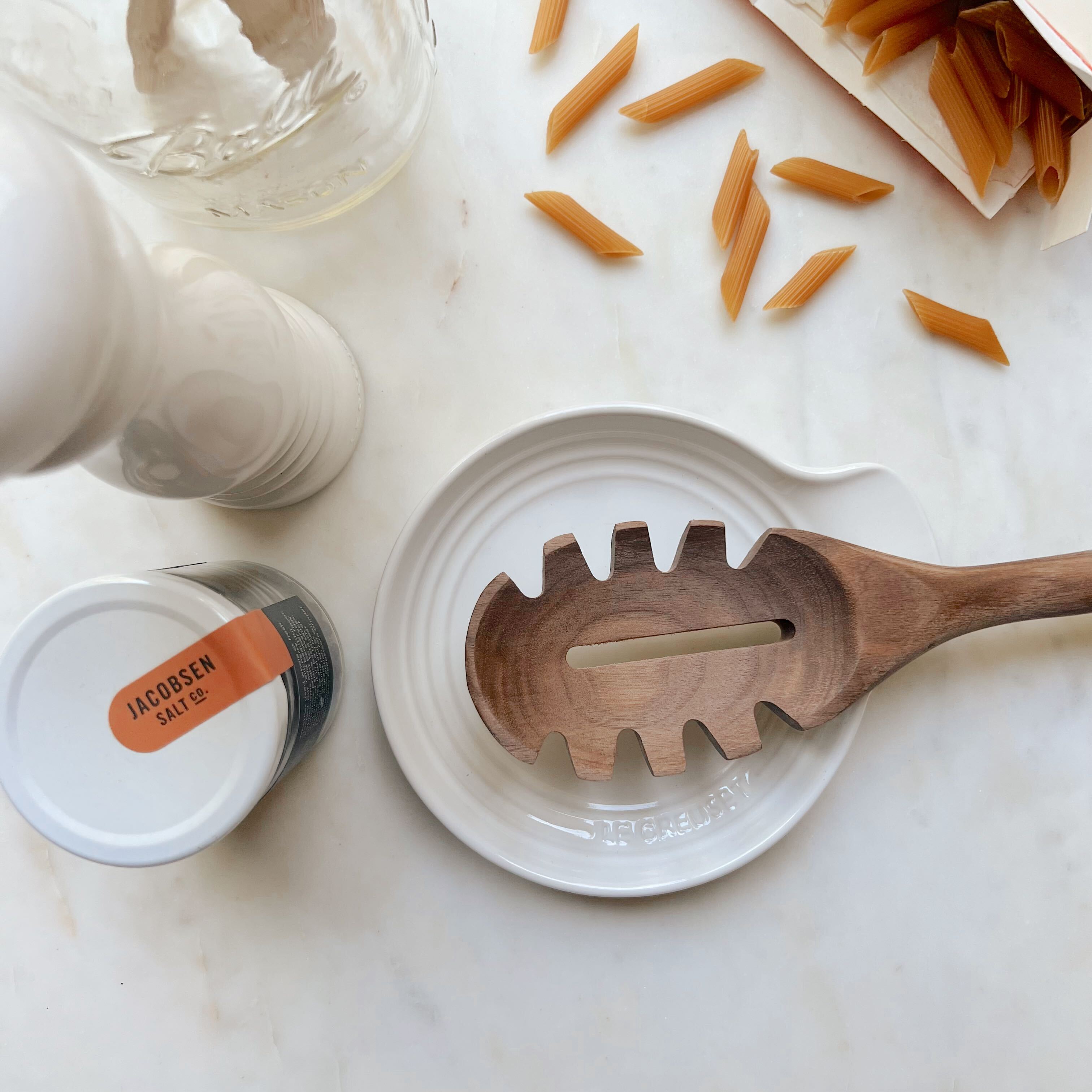 A black walnut pasta spoon laying on a white ceramic Le Creuset spoon rest next to Banza pasta noodles spilling out of their package on a white marble counter.