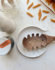 A black walnut pasta spoon laying on a white ceramic Le Creuset spoon rest next to Banza pasta noodles spilling out of their package on a white marble counter.