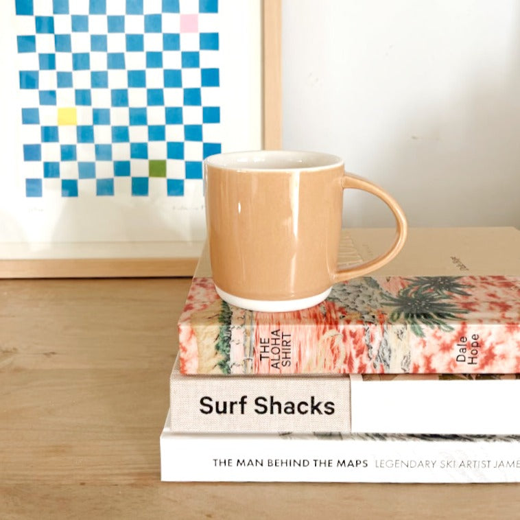 An almond shell bisque ceramic mug sitting on a stack of coffee table books on a light natural wood table in front of a blue and white checkered framed art print.
