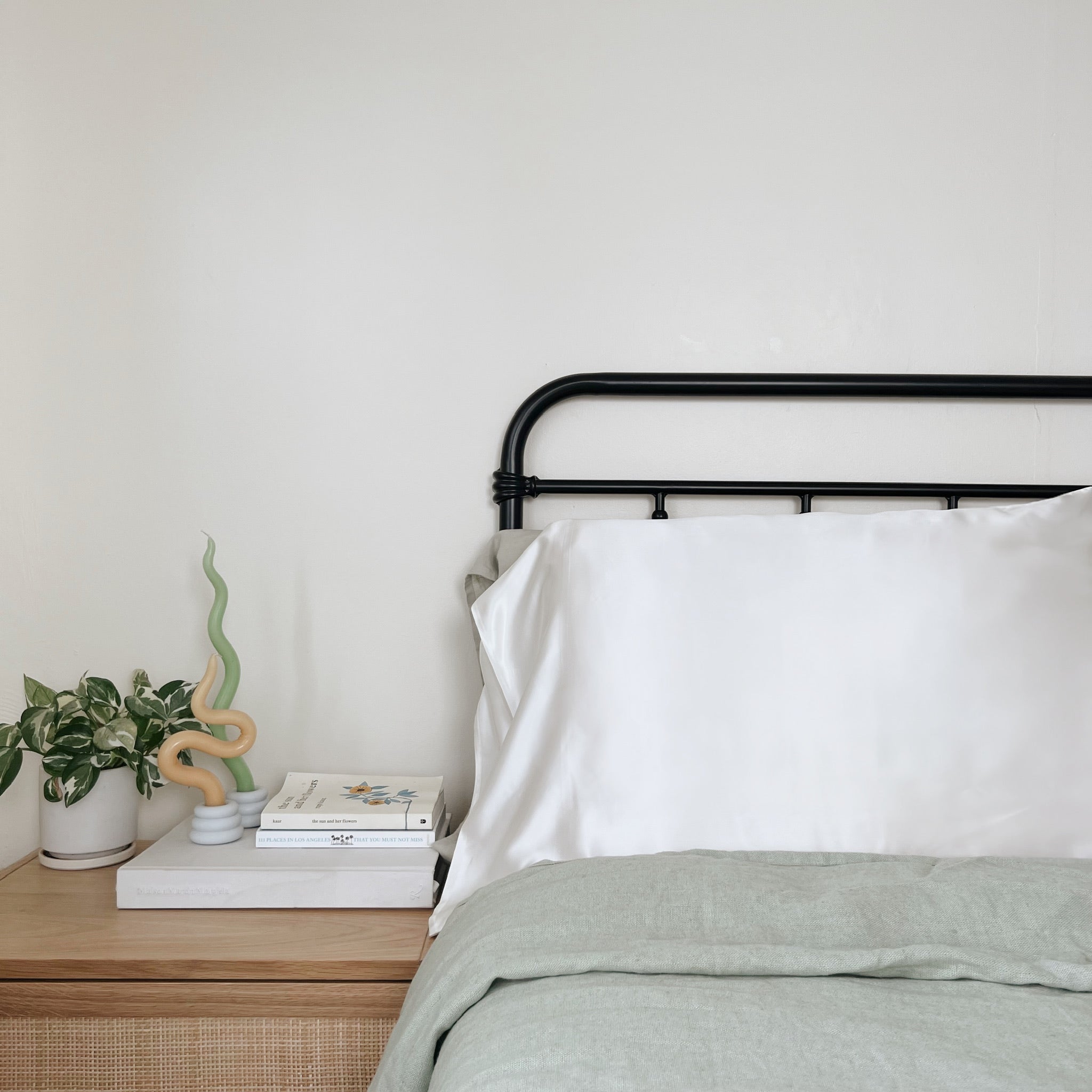 An ivory silk pillow case laying on sage green linen bedsheets on a bed with a black metal frame.