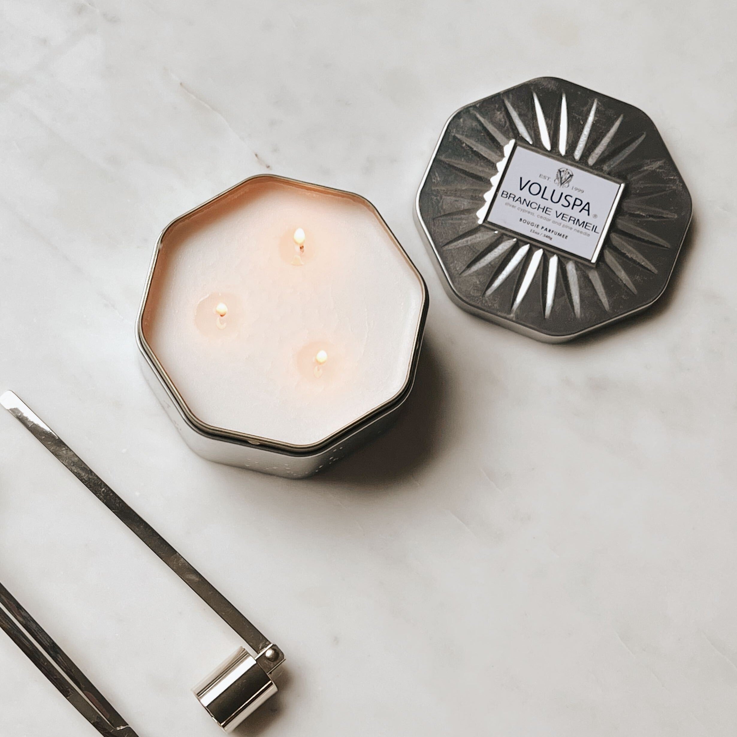 Hexagonal shaped tin with lit candle and silver candle snuffer beside it.