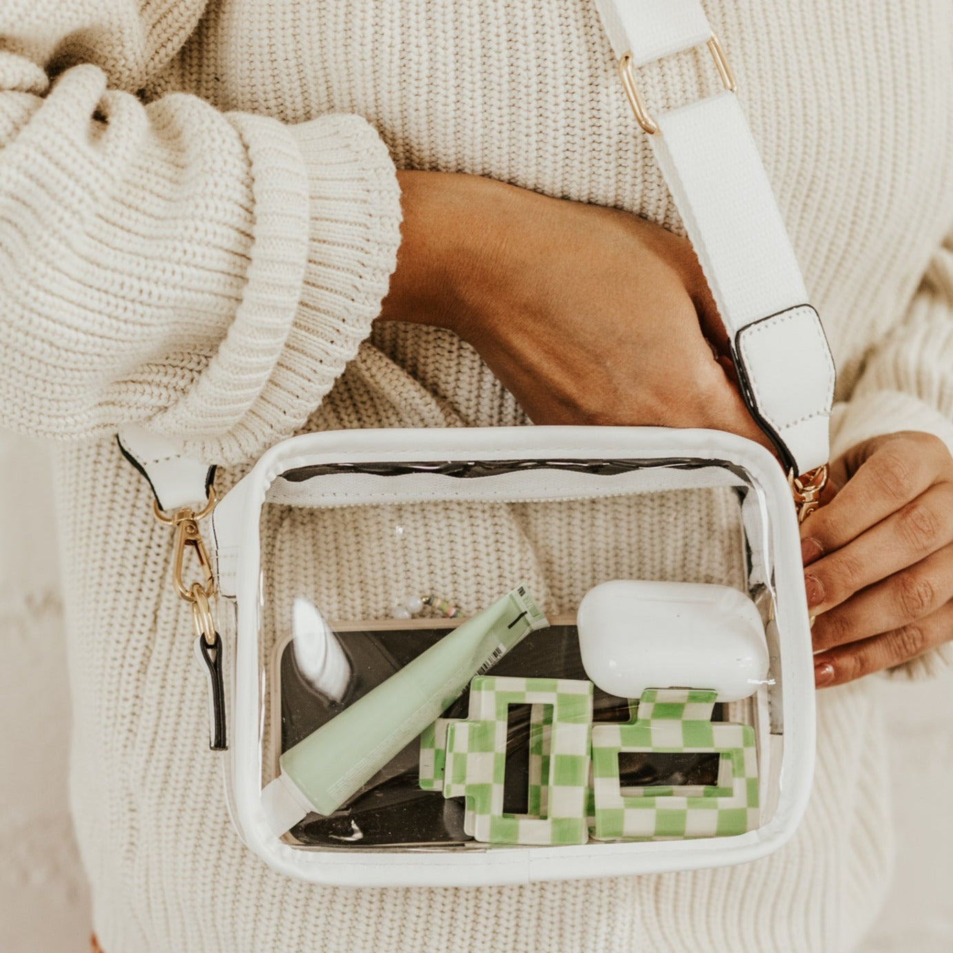 Girl wearing a cream colored sweater with a clear crossbody purse draped down her side. The purse has white lining around the edge and a white purse strap. Inside the bag is an iPhone, two small green and white checkered hair clips, a small tube of green packaged hand cream, and a set of AirPod pros.
